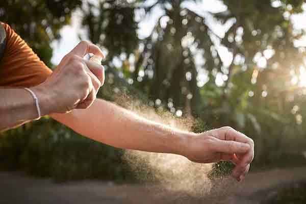 Person applying mosquito repellent on arm outdoors to prevent insect bites, with sunlight filtering through trees in the background.