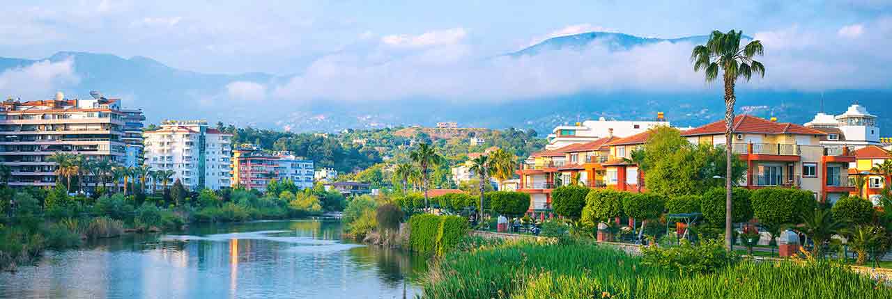 Scenic view of a vibrant residential area along a river in Turkey, showcasing modern buildings against a backdrop of lush greenery and misty mountains.