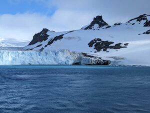  A stunning view of a glacier with icy cliffs meeting the deep blue water, surrounded by snow-capped mountains under a cloudy sky in the South Shetland Islands, Antarctica.