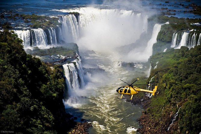 A yellow helicopter flying over the majestic Iguazu Falls with lush green forests on either side, showcasing the vast and powerful waterfalls.