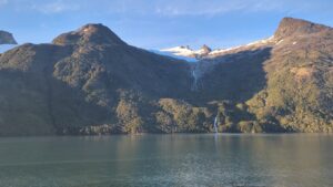 A scenic view of the Chilean Fjords with towering mountains, waterfalls cascading down rocky slopes, and calm water reflecting the landscape.